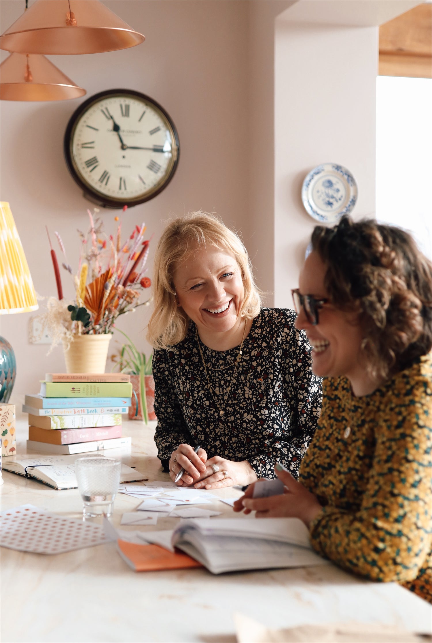 Founders Helly and Rosie, sat at a breakfast bar next to some flowers,  writing in this together cards - the perfect gift for new Mums. 